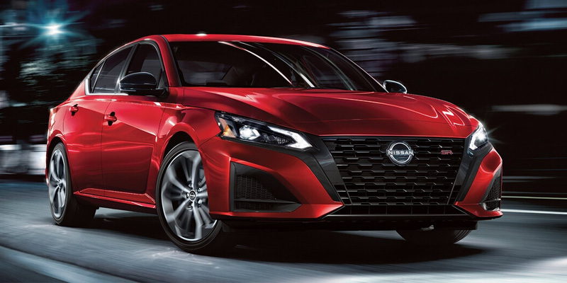 Front three-quarter view of a bright red Nissan Altima SR sedan driving down a city street at night with motion-blurred city lights in the background.